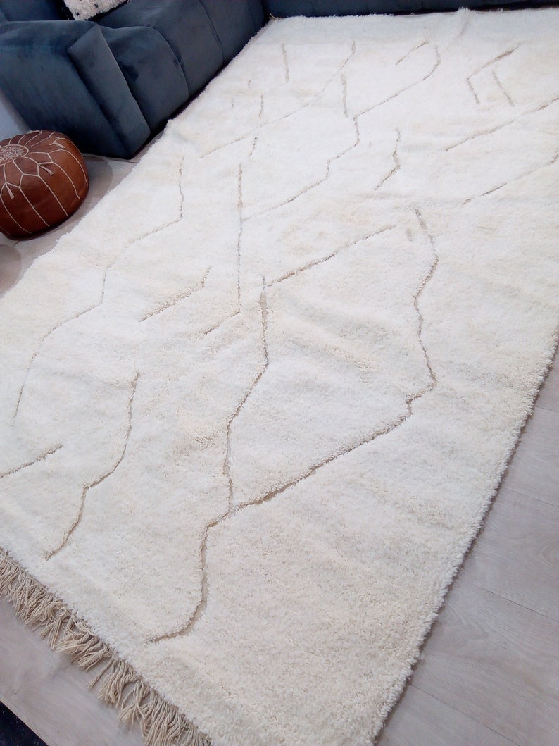 Puede incluir: Una alfombra de pelo blanco con un patr&oacute;n geom&eacute;trico en un tono de blanco m&aacute;s claro. La alfombra tiene un borde de flecos.