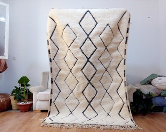 May include: A white Moroccan rug with a black diamond pattern. The rug has a fringe border and is hanging on a wall.
