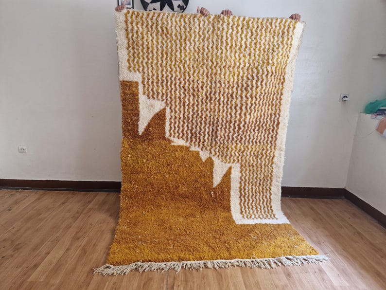May include: A large, hand-woven rug with a geometric pattern in shades of white and mustard yellow. The rug has a fringe border and is made of a soft, fluffy material.
