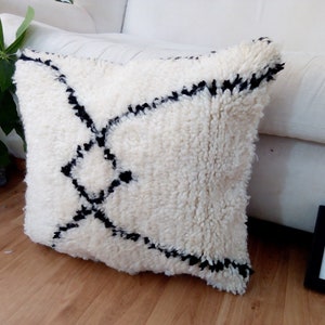 May include: White and black geometric patterned throw pillow with a fluffy texture. The pillow is on a wooden floor.