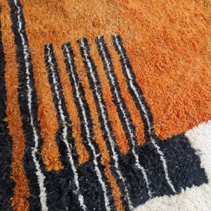 May include: A close-up of a rug with a geometric pattern in shades of orange, black, and white. The rug has a thick, fluffy texture.