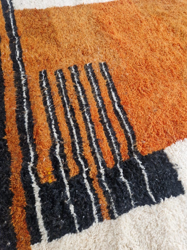 May include: A close-up of a rug with a geometric pattern in shades of orange, black, and white. The rug has a thick, fluffy texture.