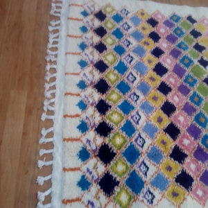 May include: A white rug with a diamond pattern in shades of blue, green, yellow, purple, and pink. The rug has fringe on the edges.