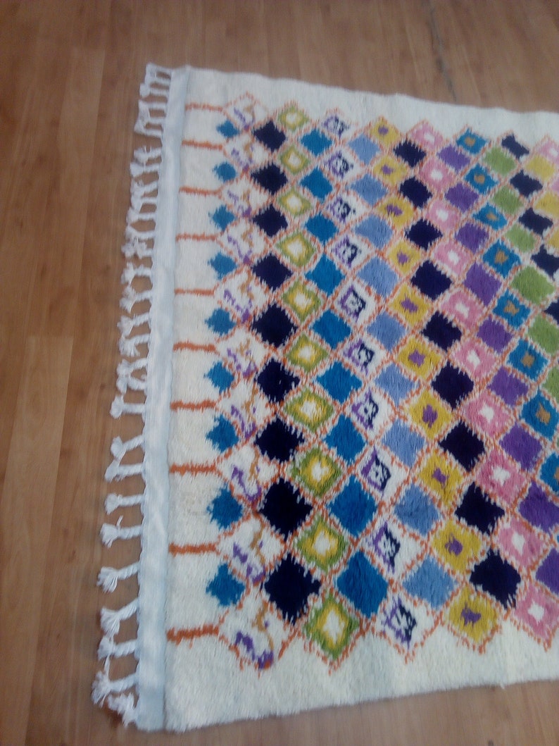 May include: A white rug with a diamond pattern in shades of blue, green, yellow, purple, and pink. The rug has fringe on the edges.