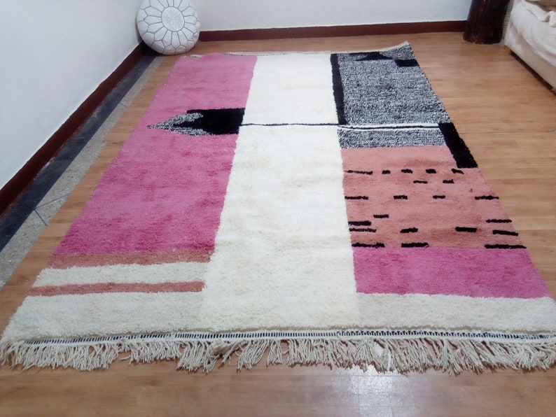 May include: A white, pink, and black geometric patterned rug with a fringe border. The rug has a modern design with abstract shapes and lines.