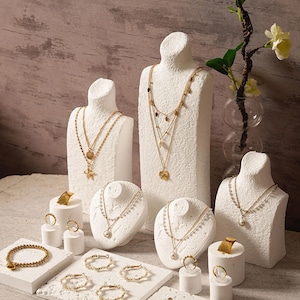 Könnte beinhalten: Eine Auswahl an goldfarbenem Schmuck, darunter Halsketten, Armbänder, Ringe und Ohrringe, präsentiert auf weißen Büsten und Sockeln. Die Halsketten weisen Anhänger und mehrschichtige Designs auf. Der Schmuck ist vor einem neutralen Hintergrund ausgestellt.