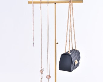 May include: Gold metal jewellery stand with a horizontal bar for hanging necklaces and a hook for a handbag. Three necklaces with pendants are hanging from the bar. A black handbag is hanging from the hook.