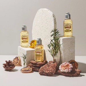 Puede incluir: Tres botellas de aceite de ducha de almendras en plataformas blancas con flores secas, piñas y rodajas de naranja. Las botellas están etiquetadas con "Amande, Huile de Douche, Shower Oil, L'Occitane."