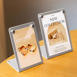 Puede incluir: Dos expositores plateados con tarjetas impresas. Una tarjeta muestra pendientes cuadrados dorados con gemas de colores. La otra tarjeta dice "NEW COLLECTION" y "CRYSTAL RINGS". Los expositores están sobre una superficie de madera.