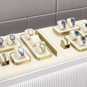 Peut inclure: Présentation de montres-bracelets variées, mettant en valeur différents styles et designs. Les montres présentent des bracelets en métal argenté et doré, avec des cadrans argentés, dorés et bleus. Elles sont présentées sur des présentoirs crème avec des bordures dorées.