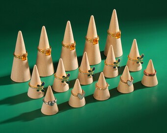 May include: A collection of rings displayed on wooden cone-shaped stands against a green background. The rings feature various designs, including gemstones, hearts, and dragonfly motifs. The rings are gold, silver, and rose gold.