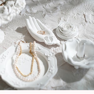 May include: White ceramic jewelry dishes in various shapes, including a hand, a rose, and a round dish with a decorative edge. The dishes are on a white textured surface. A piece of jewelry is displayed on the hand dish.