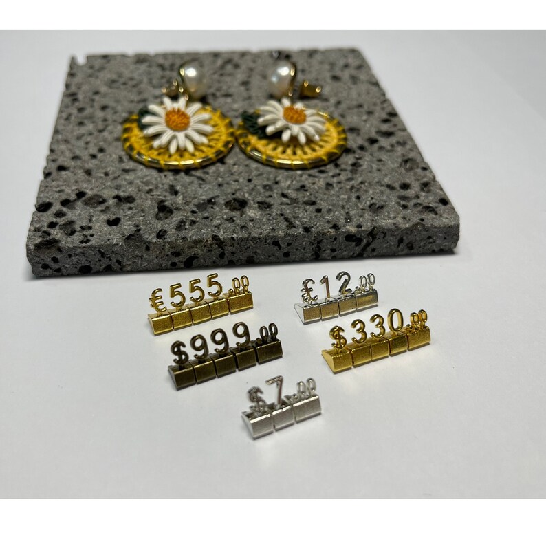 May include: A pair of yellow and white daisy earrings with pearl accents. The earrings are displayed on a gray stone surface with price tags in US dollars, Euros, and British pounds.