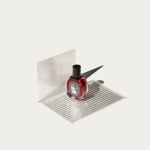 May include: A small, round, red glass bottle with a black cap sits on a clear, textured surface. The bottle has a label with a design on it.