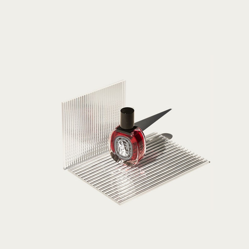 May include: A small, round, red glass bottle with a black cap sits on a clear, textured surface. The bottle has a label with a design on it.