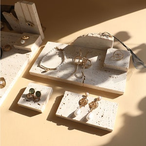May include: A set of five small, white, textured, stone-like display stands for jewelry. The stands are arranged on a beige surface. The stands feature a variety of jewelry, including a gold bracelet, a gold ring, a pair of gold earrings with pearl drops, and a pair of green stone earrings.