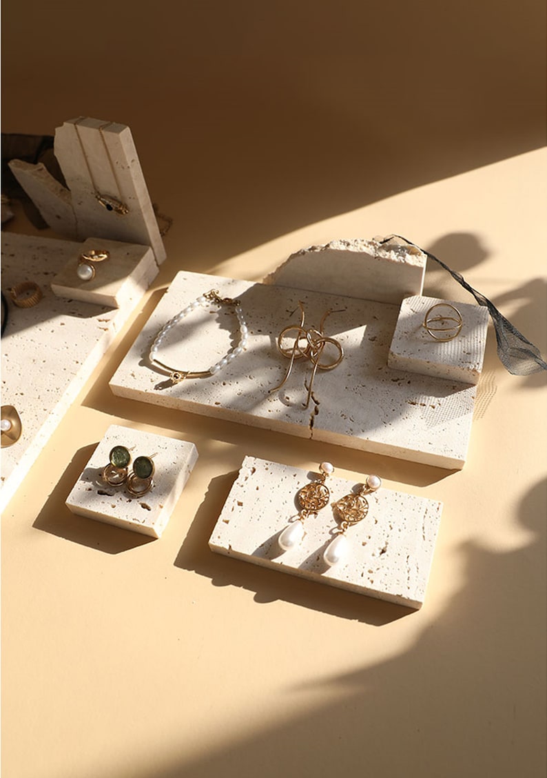 May include: A set of five small, white, textured, stone-like display stands for jewelry. The stands are arranged on a beige surface. The stands feature a variety of jewelry, including a gold bracelet, a gold ring, a pair of gold earrings with pearl drops, and a pair of green stone earrings.