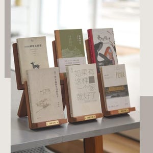 Könnte beinhalten: Fünf Bücher auf einzelnen Holzständern. Die Bücher haben verschiedene Einbanddesigns mit chinesischem Text, darunter Titel wie '如果有这样一个家就好了' und '伦敦'. Jeder Holzständer hat an der Basis ein kleines messingfarbenes Etikett.