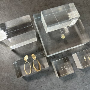 Puede incluir: Cinco expositores de acrílico transparente con diferentes piezas de joyería. Los expositores son de forma rectangular y cuadrada. La joyería incluye un collar de oro con un colgante verde, un par de aretes de aro de oro, un par de aretes ovalados de oro y un par de aretes pequeños de oro.