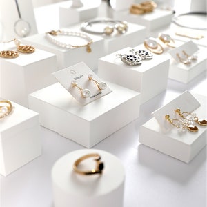 May include: White display stands with various jewelry pieces, including earrings, rings, bracelets, and necklaces. The stands are arranged on a white surface. The text "Ornaments Season Display" is printed on the surface.