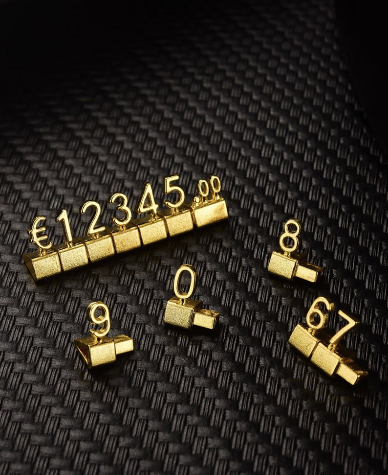 May include: Gold-colored number tiles with a euro symbol and numbers 1 through 9, 0, and 00. The tiles are arranged in a row and scattered on a black textured surface.