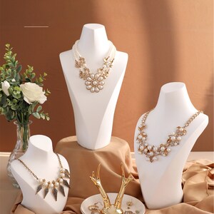 May include: Three white mannequin busts display necklaces with pearl and gold accents. A white ceramic dish with a gold deer antler ring holder sits on a brown surface.