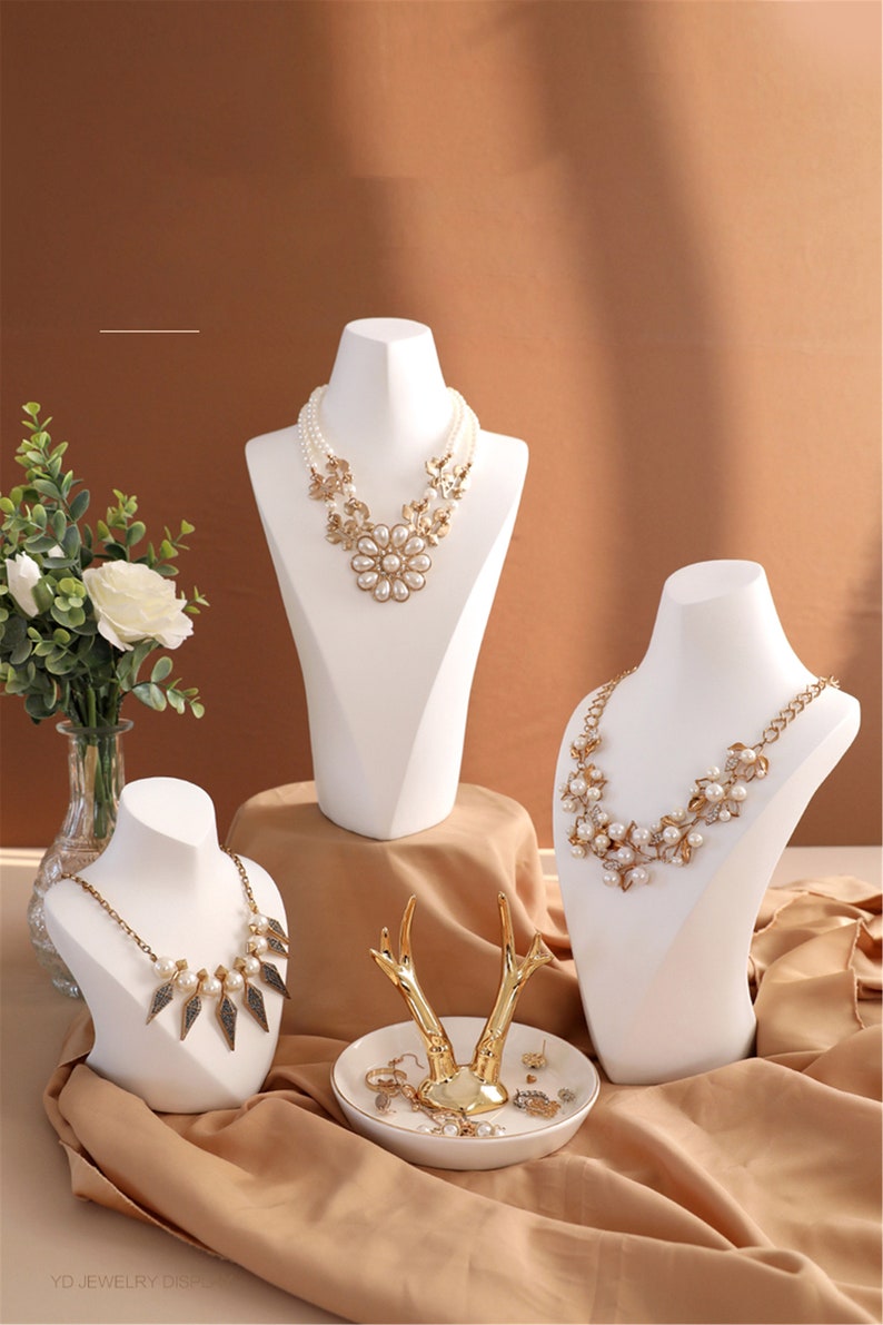 May include: Three white mannequin busts display necklaces with pearl and gold accents. A white ceramic dish with a gold deer antler ring holder sits on a brown surface.