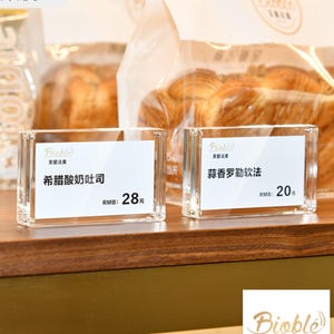 Può includere: Due porta cartelli in acrilico trasparente con carte bianche che mostrano i nomi e i prezzi degli articoli da forno. Il cartello a sinistra recita "Toast al yogurt greco" con un prezzo di 28 €. Il cartello a destra recita "Pane morbido alle erbe e all'aglio" con un prezzo di 20 €.