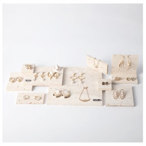 May include: A set of jewelry display stands made of beige travertine stone. The stands are arranged on a white surface and feature various gold earrings, necklaces, and rings.