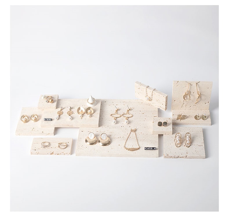 May include: A set of jewelry display stands made of beige travertine stone. The stands are arranged on a white surface and feature various gold earrings, necklaces, and rings.