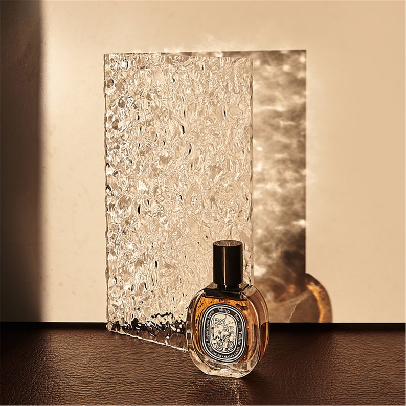 May include: A clear glass bottle of perfume with a black cap and a label that reads "Rose de Tah&iuml;ti" in a gold font. The bottle is sitting on a textured surface.