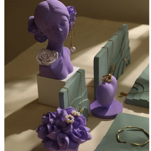 May include: Purple jewelry display stands with a bust of a woman with a flower in her hair, a heart-shaped stand, and a rectangular stand with a groove for necklaces. The stands are made of a smooth, matte material.
