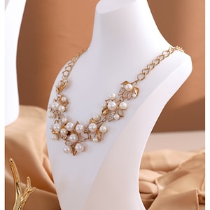 May include: A gold necklace with a floral design and white pearls on a white mannequin bust. The necklace is on a light brown fabric background.