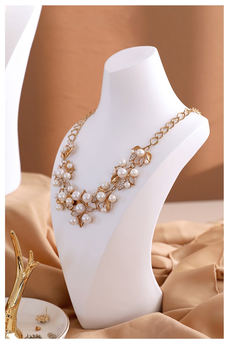 May include: A gold necklace with a floral design and white pearls on a white mannequin bust. The necklace is on a light brown fabric background.