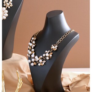 May include: A black jewellery display stand with a gold and pearl necklace. The necklace features a floral design with small white pearls and gold accents.