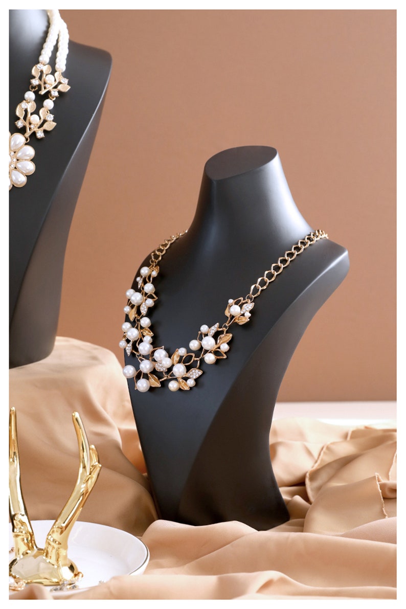 May include: A black jewellery display stand with a gold and pearl necklace. The necklace features a floral design with small white pearls and gold accents.