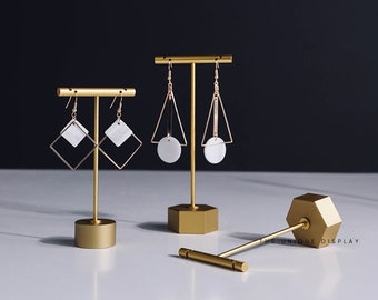 Può includere: Tre espositori per orecchini in metallo dorato. Gli espositori sono a forma di esagono, cerchio e quadrato. Gli espositori sono su una superficie bianca. Il testo "THE UNIQUE DISPLAY" è visibile sullo stand a forma di esagono.