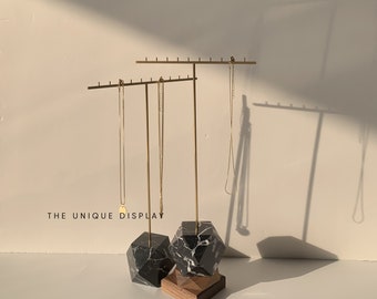 May include: A gold metal jewellery stand with two bars for hanging necklaces. The stand has a black and white marble base with a wooden platform. The text "THE UNIQUE DISPLAY" is visible in the image.