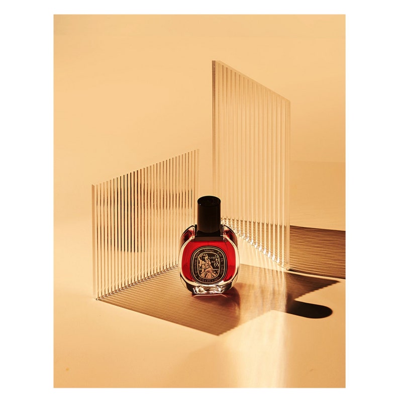 May include: A dark red glass bottle of perfume with a black cap. The bottle is sitting on a white surface with a clear acrylic background.