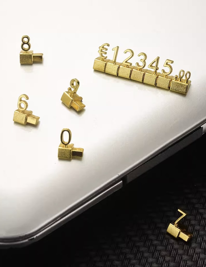 May include: Gold-colored number cubes with the numbers 1 through 5, a euro symbol, and a 00. The cubes are arranged in a row. There are also individual gold-colored number cubes with the numbers 6, 7, 8, 9, and 0.