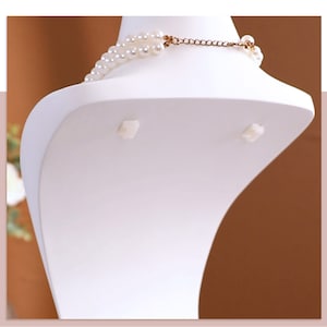 May include: White plastic bust form for displaying necklaces. The bust has a curved shape and two small white tabs on the back.