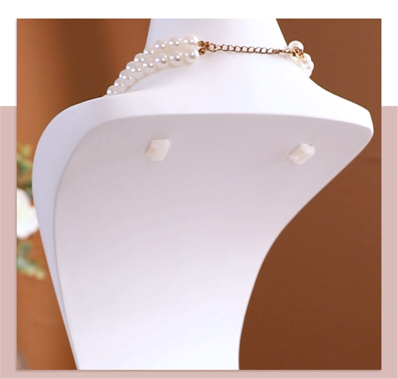 May include: White plastic bust form for displaying necklaces. The bust has a curved shape and two small white tabs on the back.