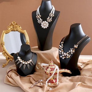 May include: Three black jewellery display busts with pearl necklaces. The necklaces feature different styles of pearl and metal accents. A gold genie lamp with a pearl necklace draped over it sits in front of the busts.