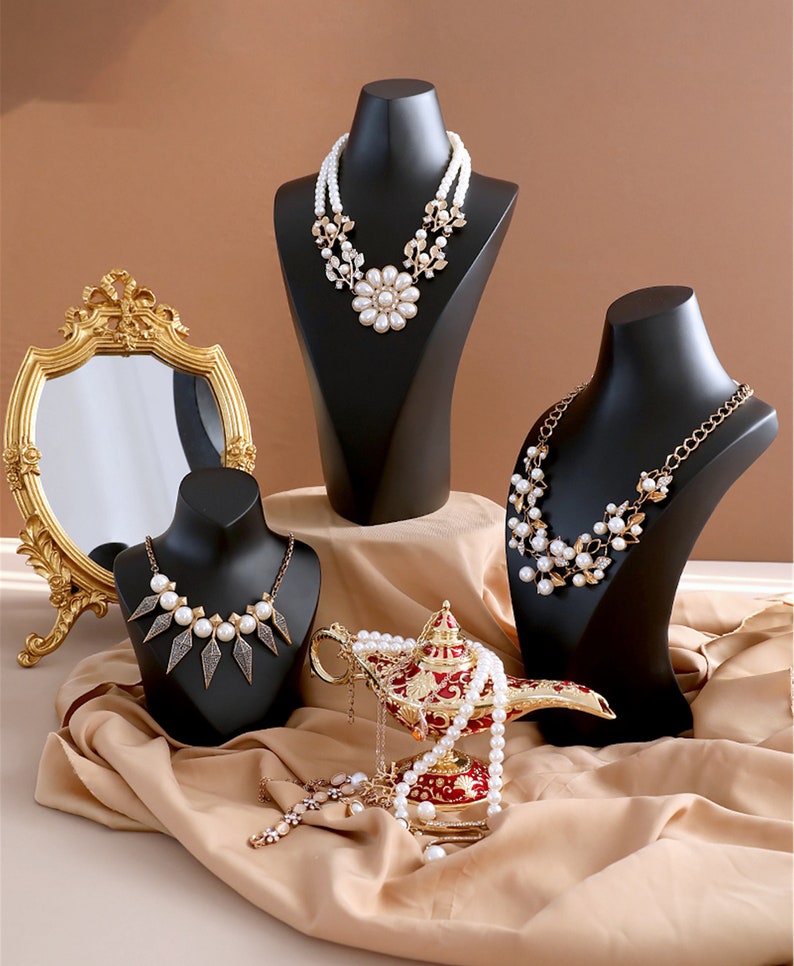 May include: Three black jewellery display busts with pearl necklaces. The necklaces feature different styles of pearl and metal accents. A gold genie lamp with a pearl necklace draped over it sits in front of the busts.
