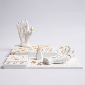 May include: White ceramic jewelry display stand with multiple hands and a cone shaped ring holder. The stand is decorated with small white beads. Gold chains, bracelets, and rings are displayed on the stand.