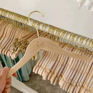 Könnte beinhalten: Kleiderbügel aus Holz mit goldfarbenen Metallhaken. Die Kleiderbügel sind hellbraun mit einem Schwan-Logo und dem Text "LOUIS MIND". Die Kleiderbügel werden an einer goldfarbenen Metallstange präsentiert.