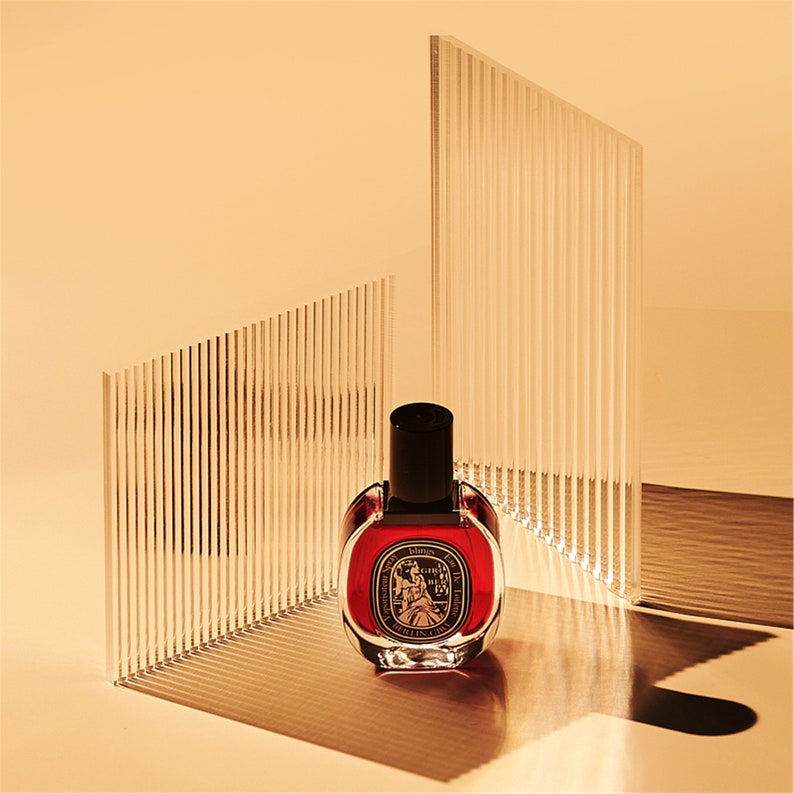 May include: A red glass bottle of perfume with a black cap and gold accents. The bottle is sitting on a white surface with a striped background.