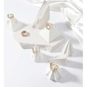 May include: White ceramic ring display stands in various shapes, including pyramids and cones, with gold rings on top. The stands are on a white background.