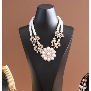 May include: A white pearl necklace with a gold flower pendant and leaf accents. The necklace is displayed on a black jewellery stand.
