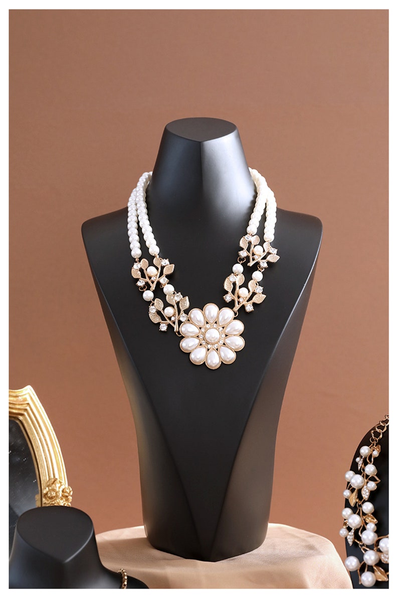 May include: A white pearl necklace with a gold flower pendant and leaf accents. The necklace is displayed on a black jewellery stand.